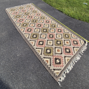 Full high-angle shot of a vintage 4'x9' Balkan tribal Kilim runner rug on asphalt, showcasing repeating terracotta, pink, and olive stepped diamonds.